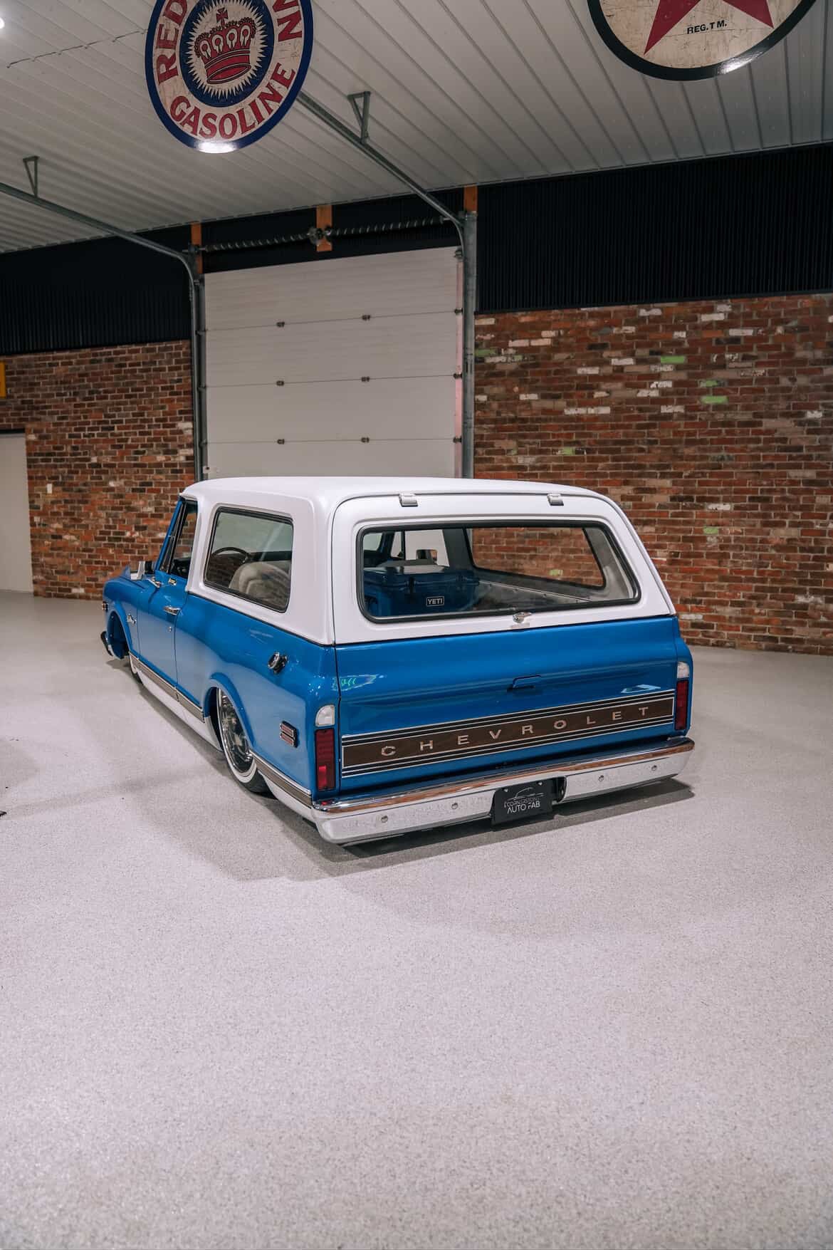 Vintage blue and white Chevrolet truck in a garage with a brick wall and 'Chevy' sign.