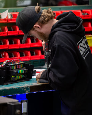 Person wearing a black hoodie with a logo, working on a task at a table with red storage bins in the background.