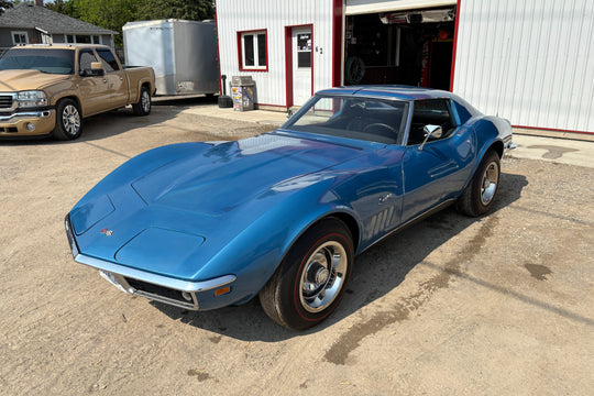 Blue vintage car parked in front of a building with a truck and trailer in the background.