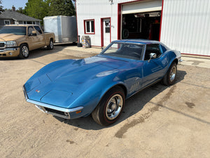 Blue vintage 1969 Corvette parked in front of a building with a truck and trailer in the background.
