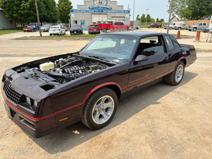 Black muscle car with hood open on a street with buildings and cars in the background