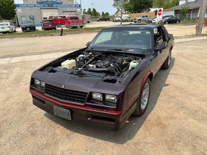 Vintage car with its hood open on a dirt road, with a gas station and vehicles in the background.