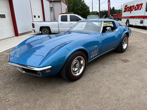 Blue vintage car parked on a gravel driveway with a building and truck in the background.