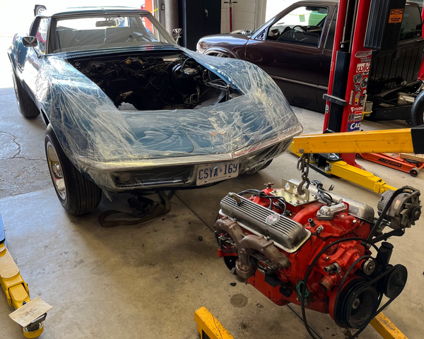 Car with its hood open in a garage, next to a red engine on a lift.